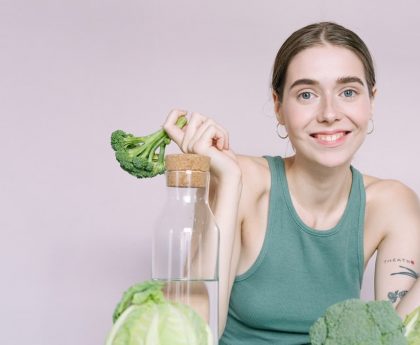 Woman in tank top smiling with broccoli, cabbage, and glass of water on table.