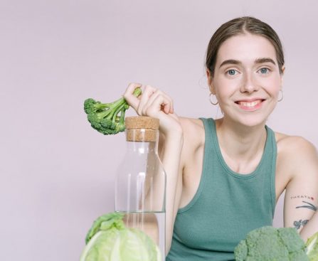 Woman in tank top smiling with broccoli, cabbage, and glass of water on table.
