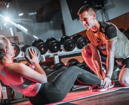 A personal trainer assists a woman doing sit-ups with a medicine ball in a gym setting.